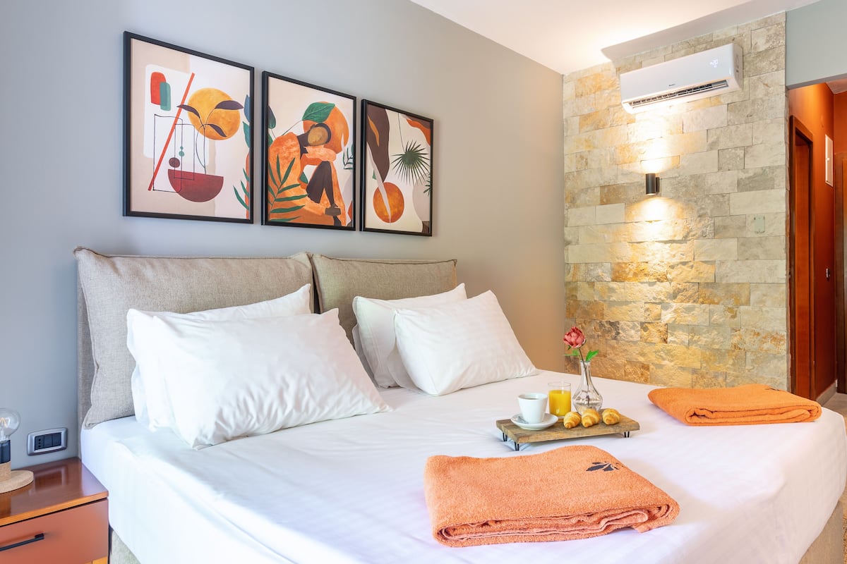 A hotel room with a bed, white linens, a tray with a drink and fruit, a vase with a flower, and three framed artworks on the wall.