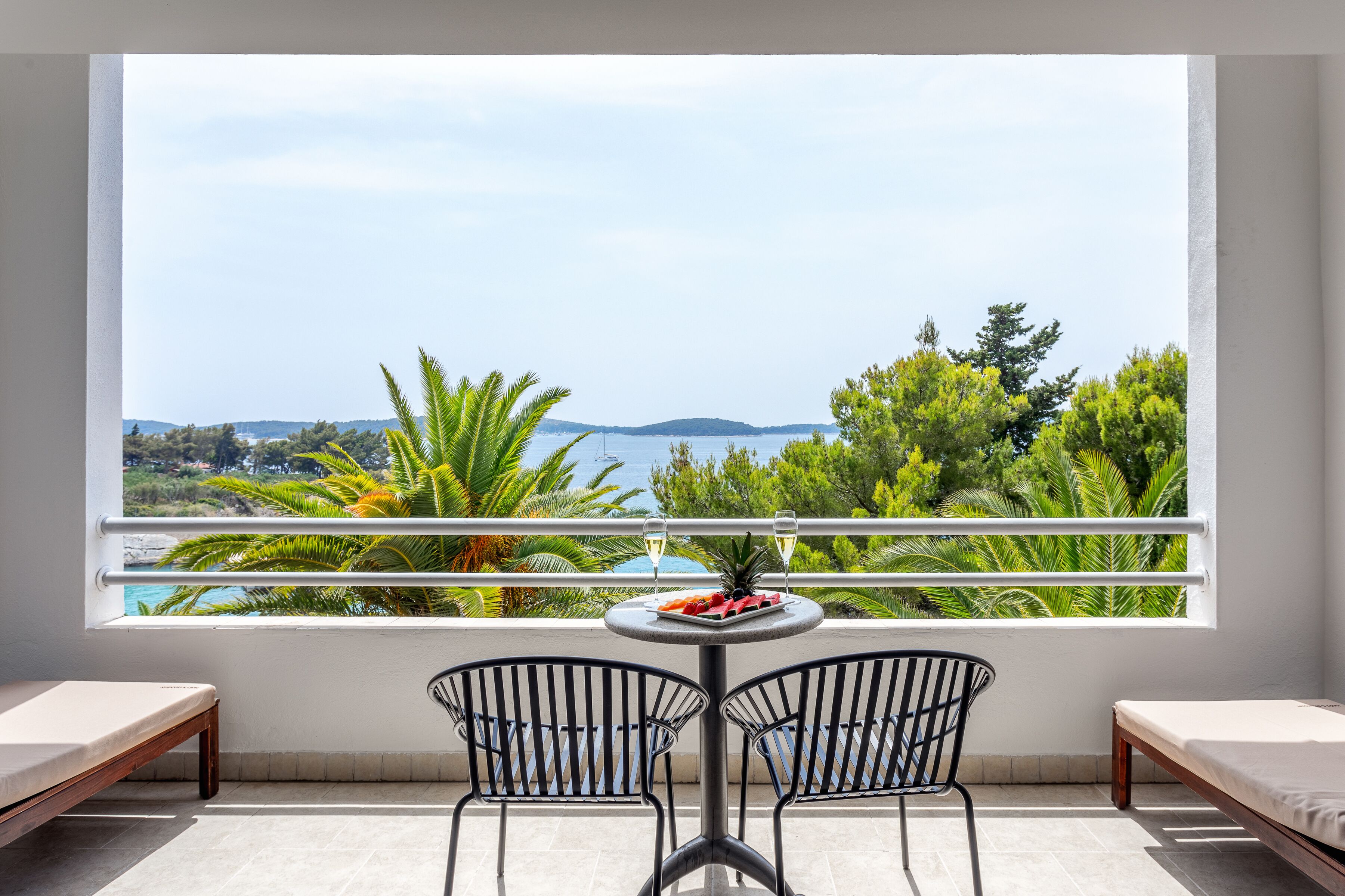 Een balkon met uitzicht op zee, twee stoelen, een tafel met een schaal fruit en een fles wijn.