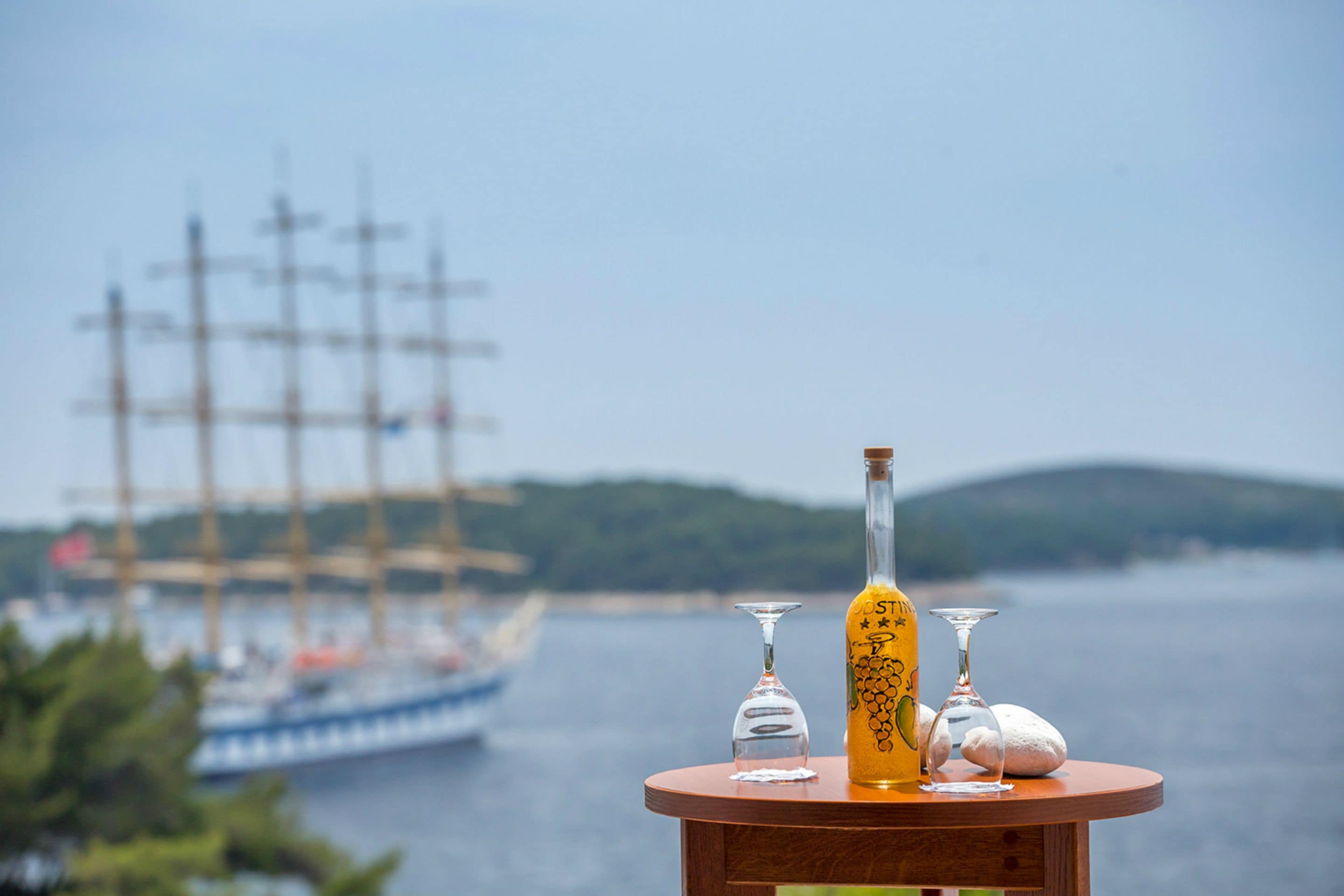 A table with a bottle and two glasses, overlooking a body of water with a ship in the distance.