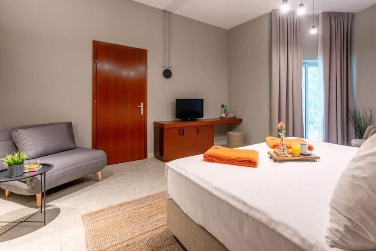 A modern hotel room with a grey sofa, a bed with an orange towel, a TV on a wooden cabinet, and a window with curtains.