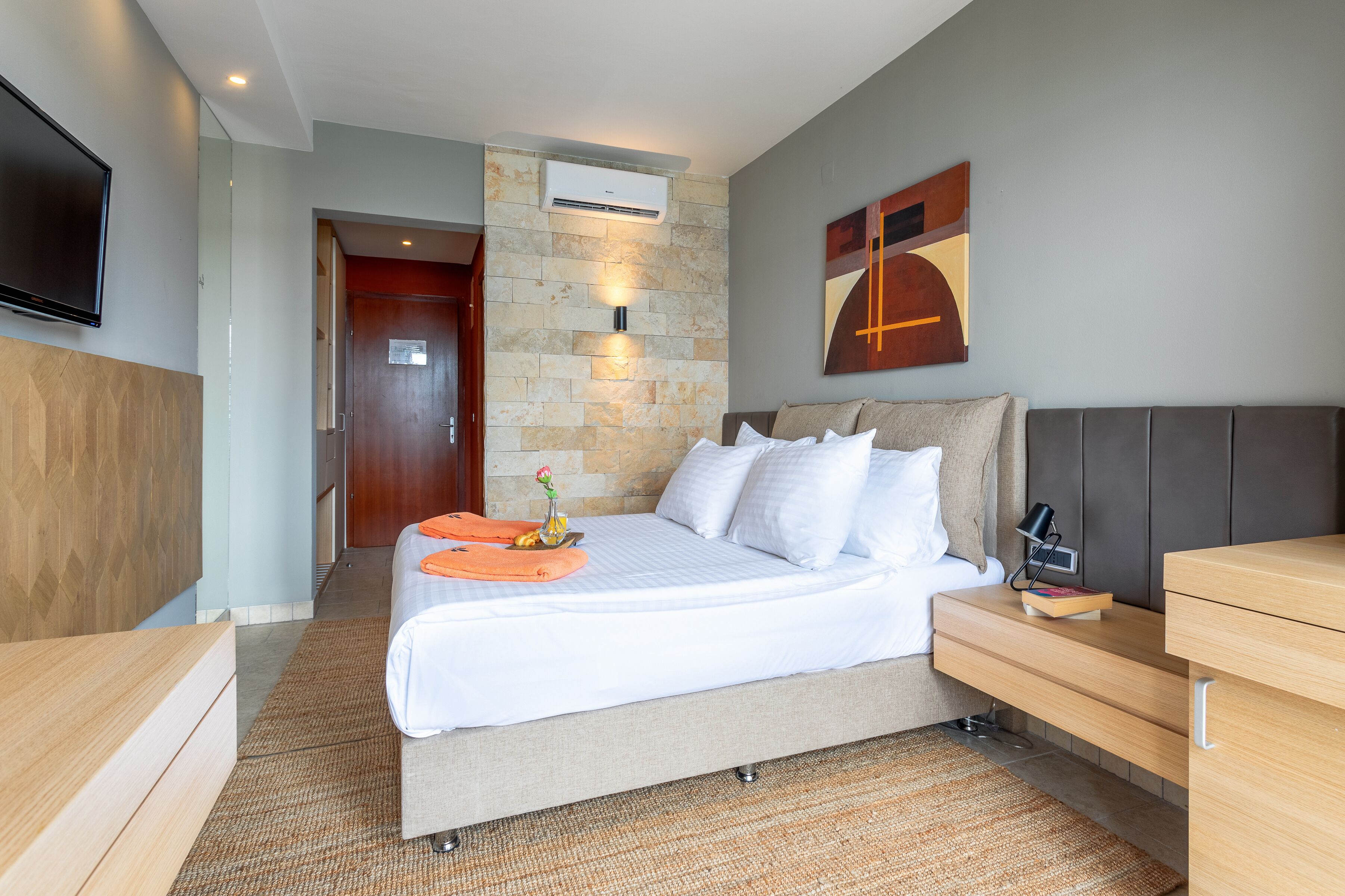 A modern hotel room with a large bed, a desk, a flat-screen TV, and a stone accent wall.