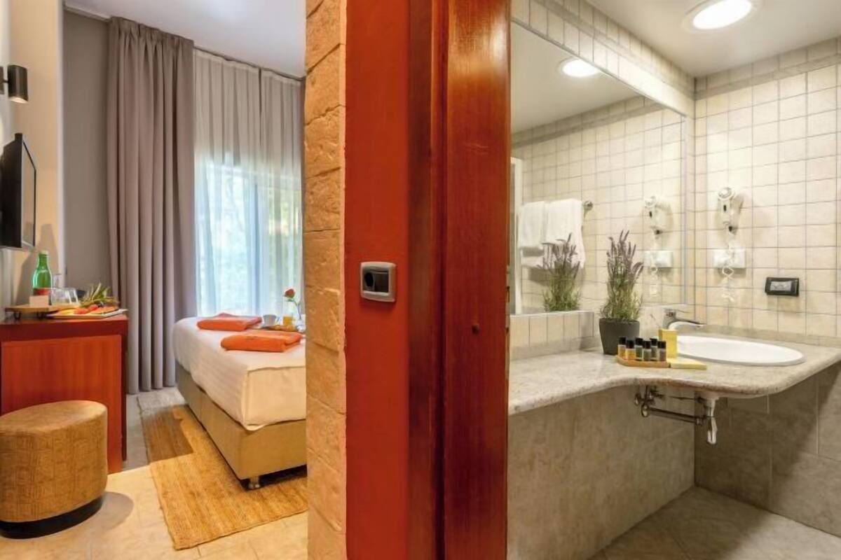 A hotel room with a bed, a wooden desk, a small stool, a bathroom with a sink, and a mirror.