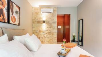Habitación de hotel con cama, almohadas, una bandeja con una flor, una taza y un plato de comida. Se observa una pared de piedra, un equipo de aire acondicionado y una puerta de madera.
