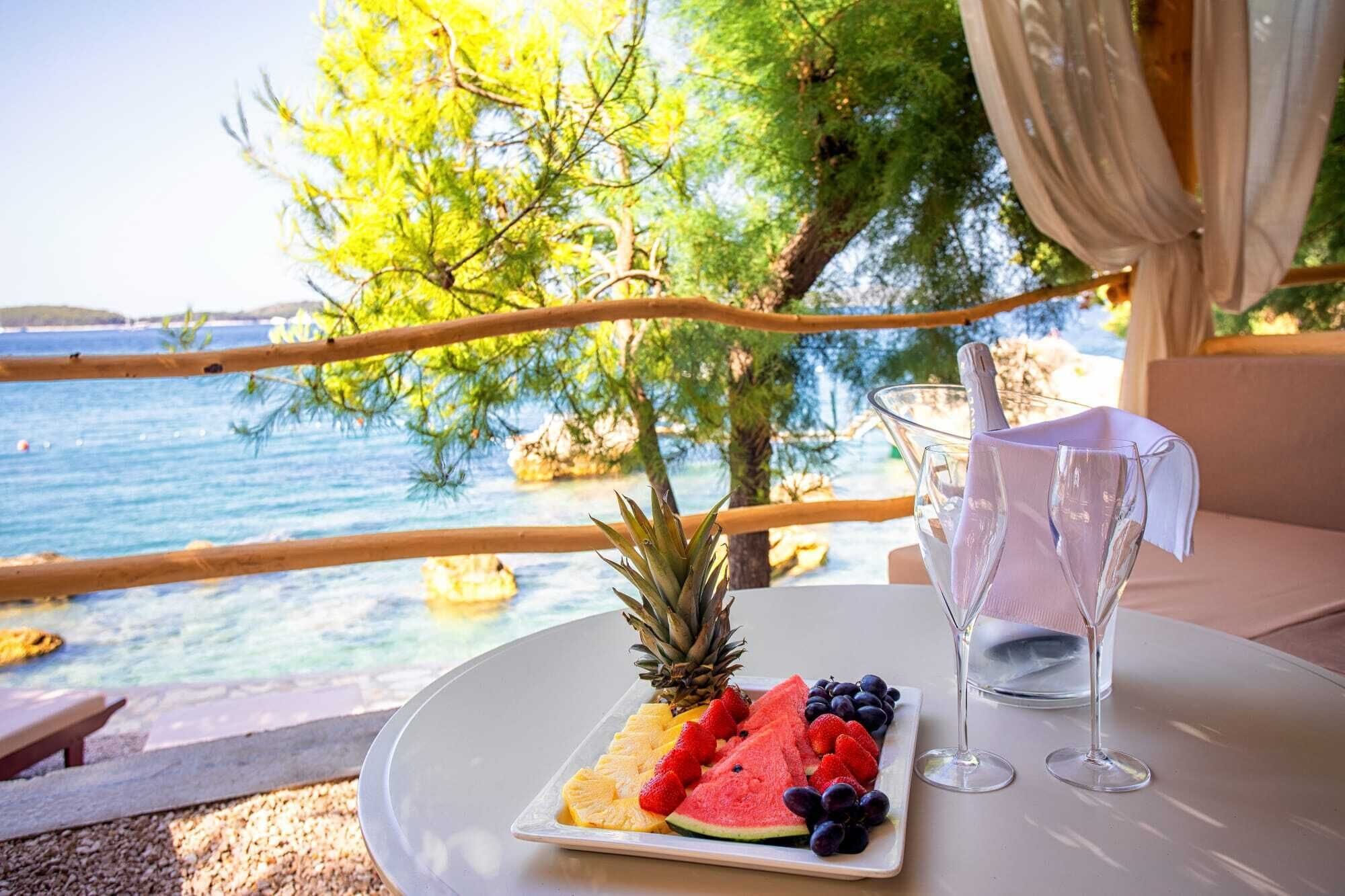 Una mesa con una bandeja de frutas, dos copas de vino y vistas al mar.