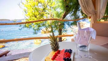 A table with a fruit platter, two wine glasses, and a view of the sea.