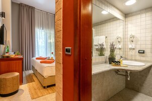 Una habitación de hotel con cama, mesita de noche y baño con lavamanos y toallero.