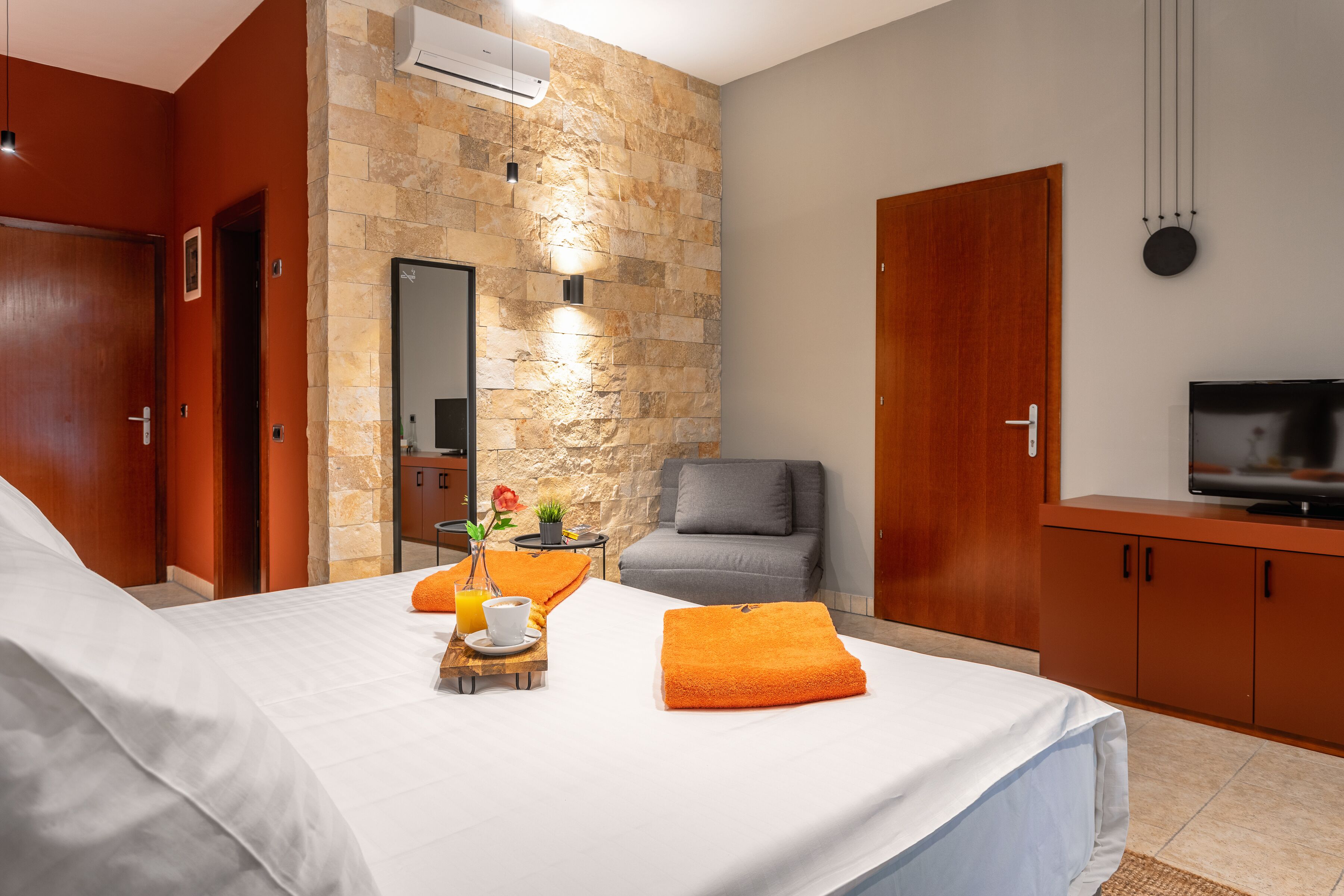 A hotel room with a bed, a desk with a chair, a TV, and a stone wall.