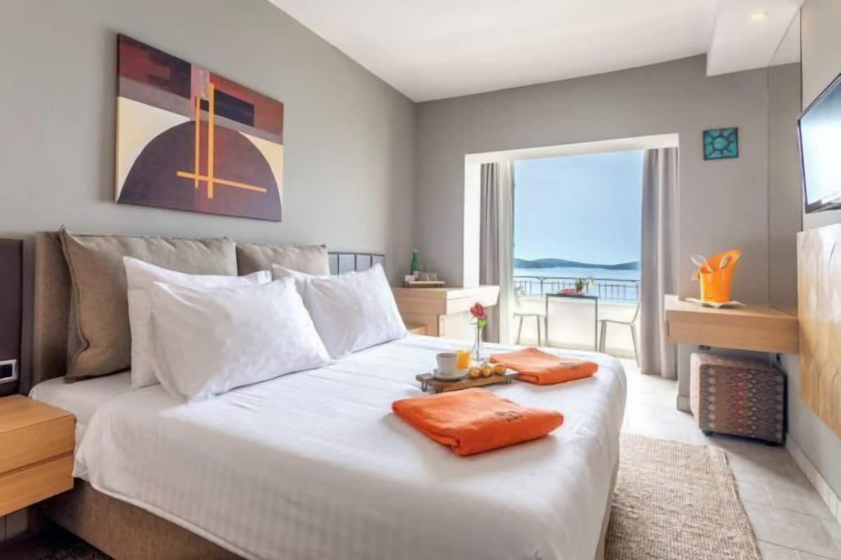A hotel room with a bed, a bedside table, a nightstand, a window with a view, and a painting on the wall.