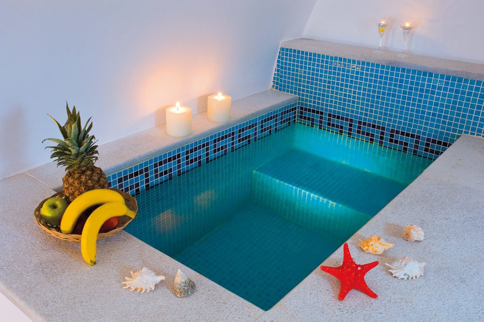 Una pequeña piscina con azulejos de mosaico azul, rodeada por una terraza de piedra. Hay velas en la pared y una cesta de frutas con piña, manzana y plátanos.
