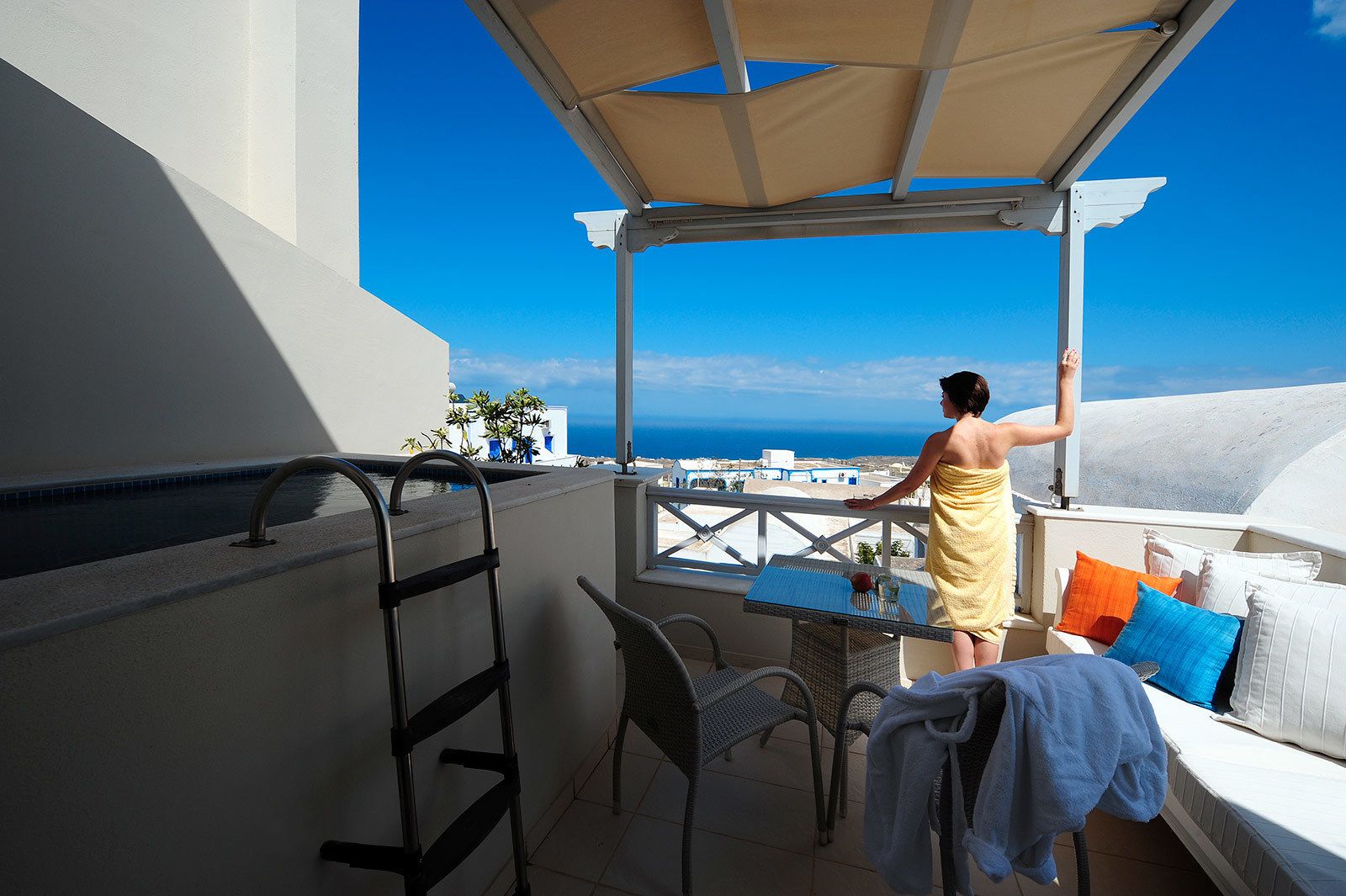 A balcony with a view of the sea, a table with a glass, and a person standing under a canopy.