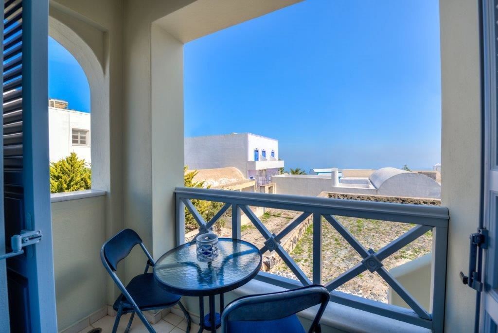 A balcony with a table and chairs, overlooking a coastal area with white buildings and clear blue sky.