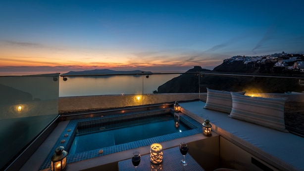 A rooftop infinity pool with a view of the sunset over the sea, a cozy seating area with cushions, and a glass balustrade.