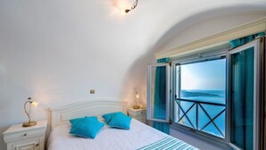A bedroom with a bed, two nightstands, a window with curtains, and a view of the sea.
