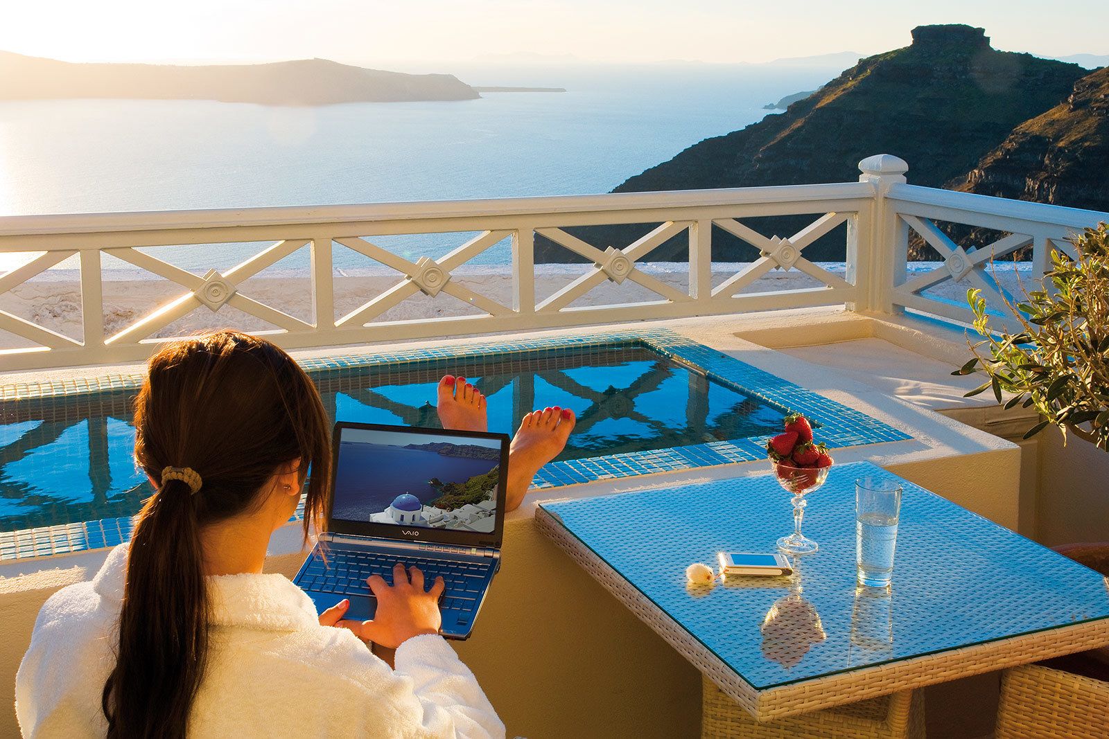 A person using a laptop at a poolside table with a glass of water and a fruit garnish, overlooking a scenic coastal view.