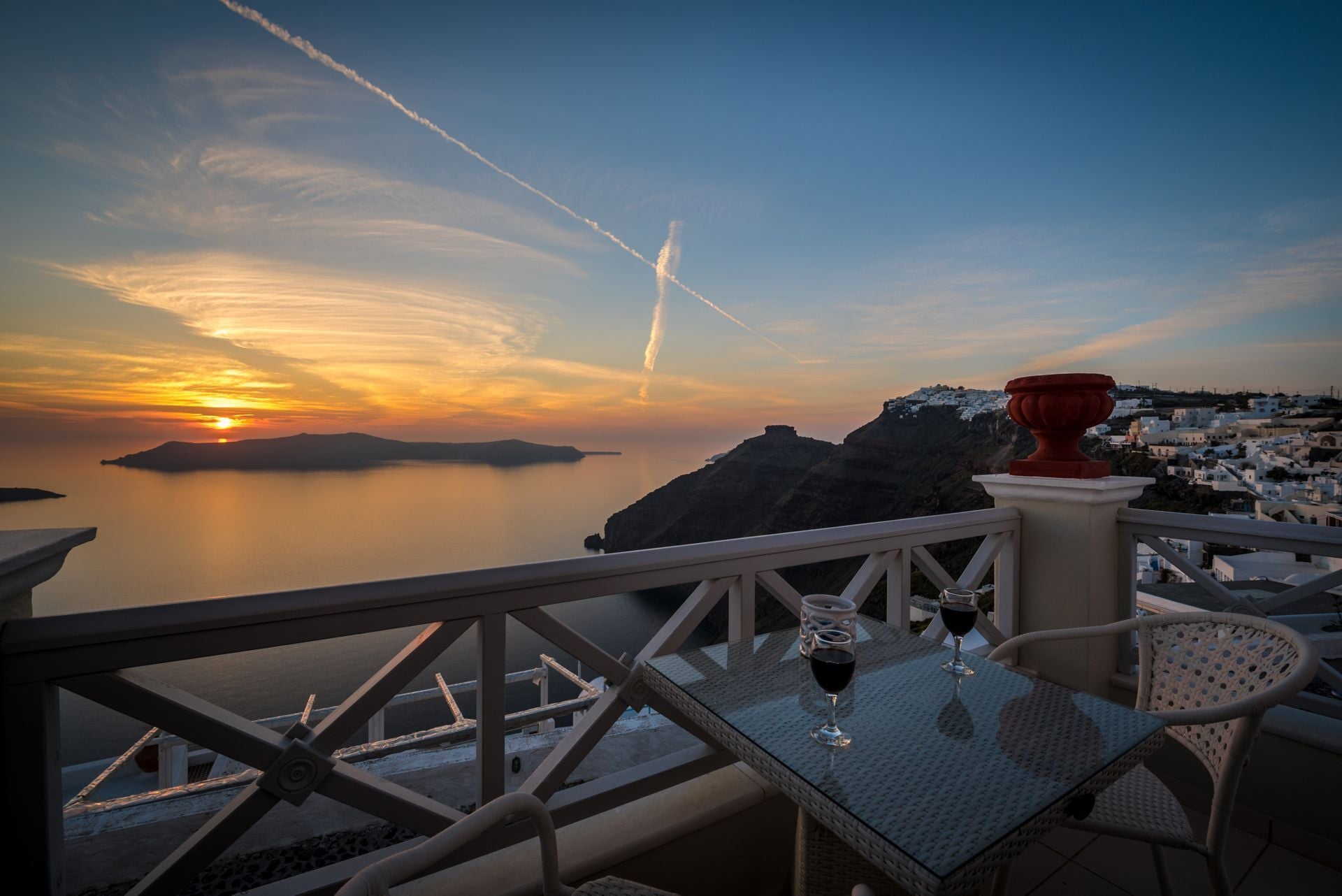 Un balcón con mesa y sillas, con vista a un paisaje costero al atardecer y un cielo despejado.