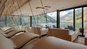 Una habitación de hotel moderna con una cama grande, zona de estar y vistas panorámicas a la montaña y al lago.