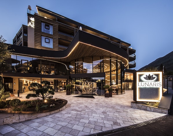 A modern hotel with a curved glass facade, outdoor seating area, and a sign reading "LUNARIS". - LUNARIS Wellnessresort (Valle Aurina)