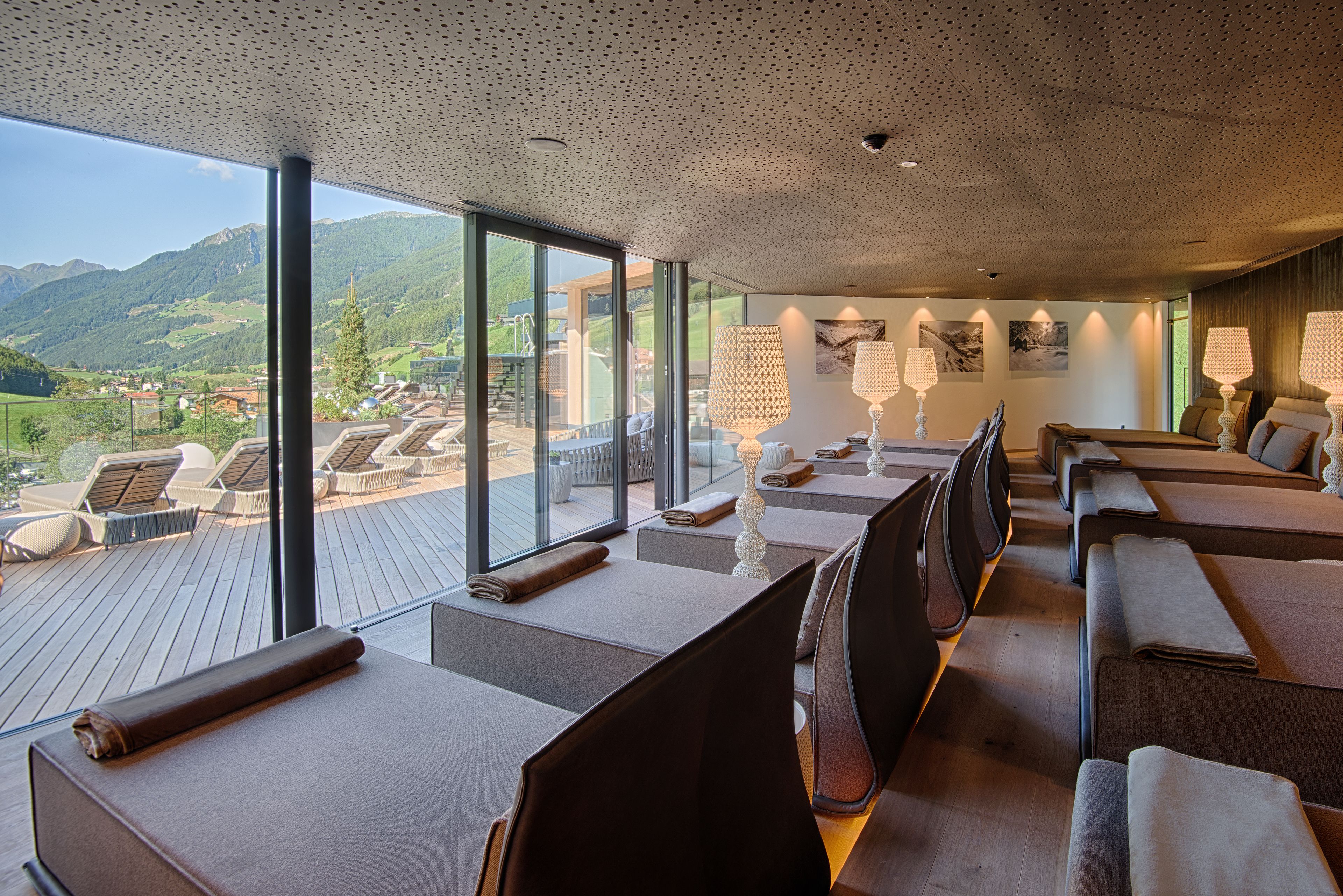 Ein moderner Wellnessbereich mit großen Fenstern und Blick auf die Berge. Mehrere Massagetische sind mit Handtüchern ausgestattet, außerdem steht eine große Stehlampe bereit.