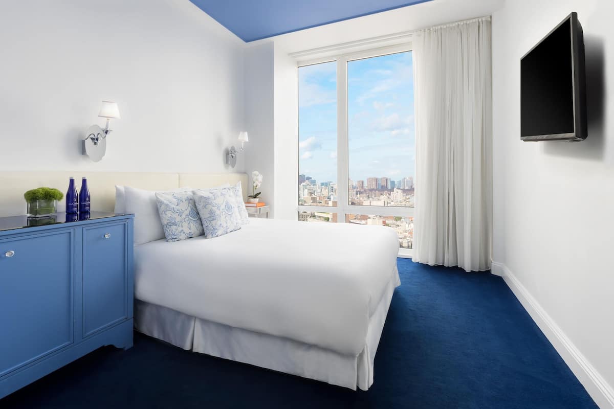 Une chambre d’hôtel avec un grand lit, une commode bleue, un téléviseur à écran plat et une vue sur la ville par la fenêtre.