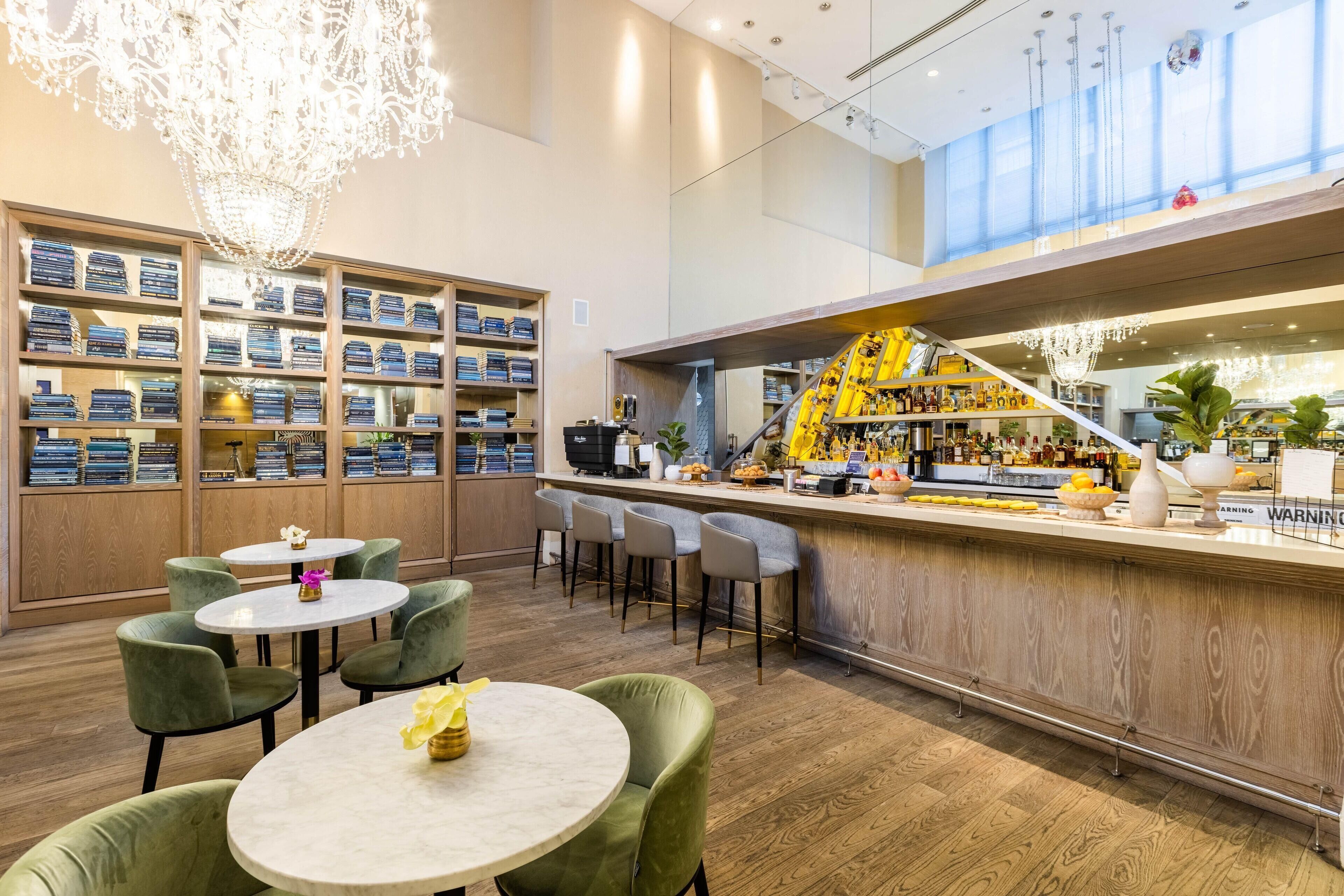 A bar area with a wooden counter, bar stools, and a chandelier. There are shelves with bottles and a small table with a vase.