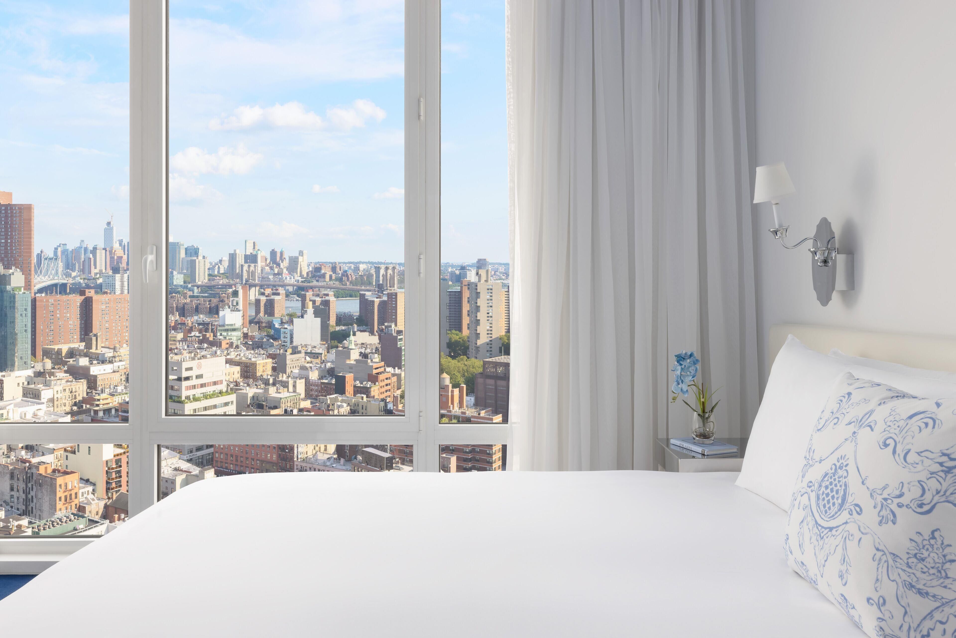 A hotel room with a large window offering a city view, a bed with white bedding, a nightstand with a lamp, and a vase with flowers.
