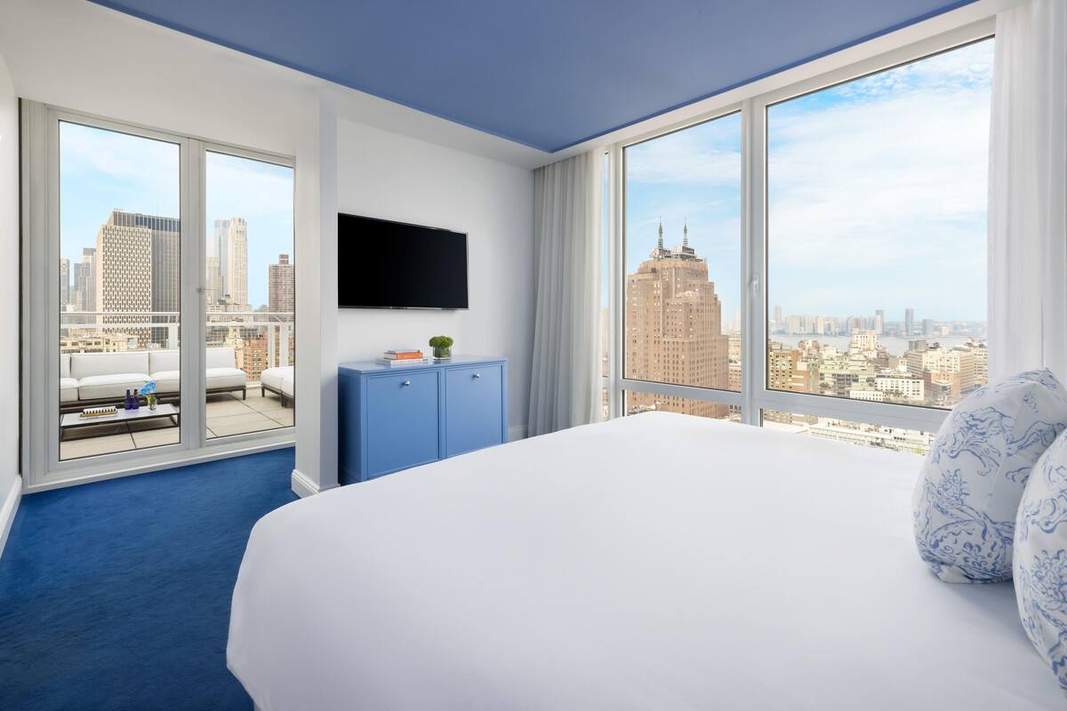 Une chambre d’hôtel avec un grand lit, un téléviseur à écran plat, une armoire bleue et une vue sur la ville à travers de grandes fenêtres.