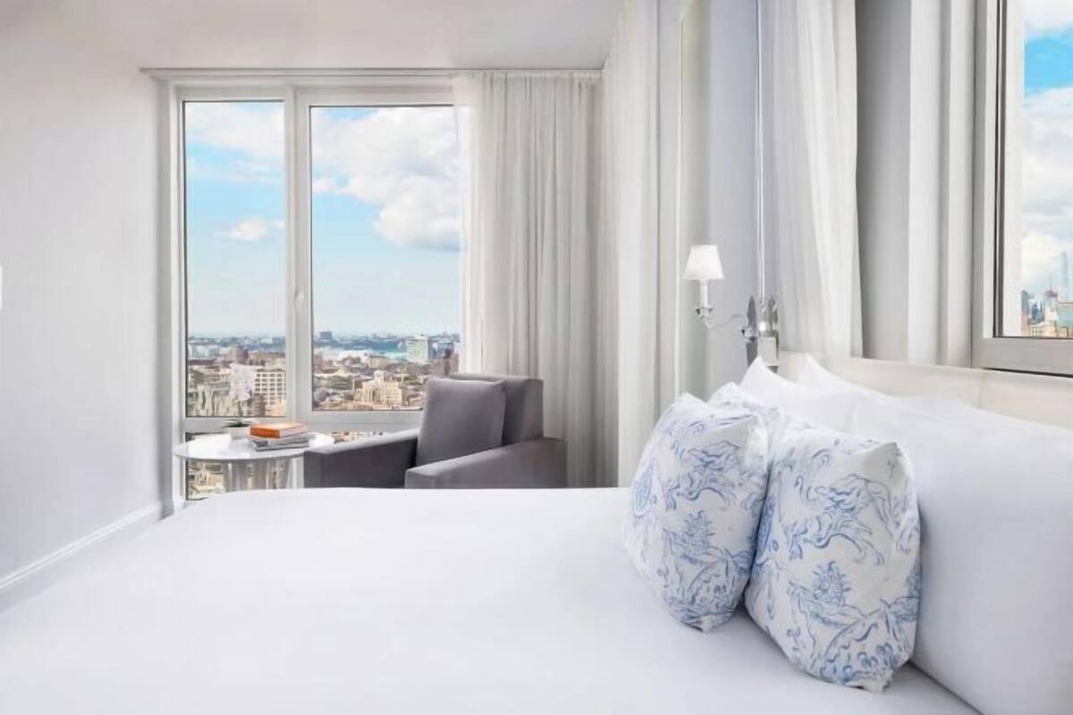 Une chambre d’hôtel avec un grand lit, une chaise, une petite table et une vue sur le paysage urbain grâce à de grandes fenêtres.