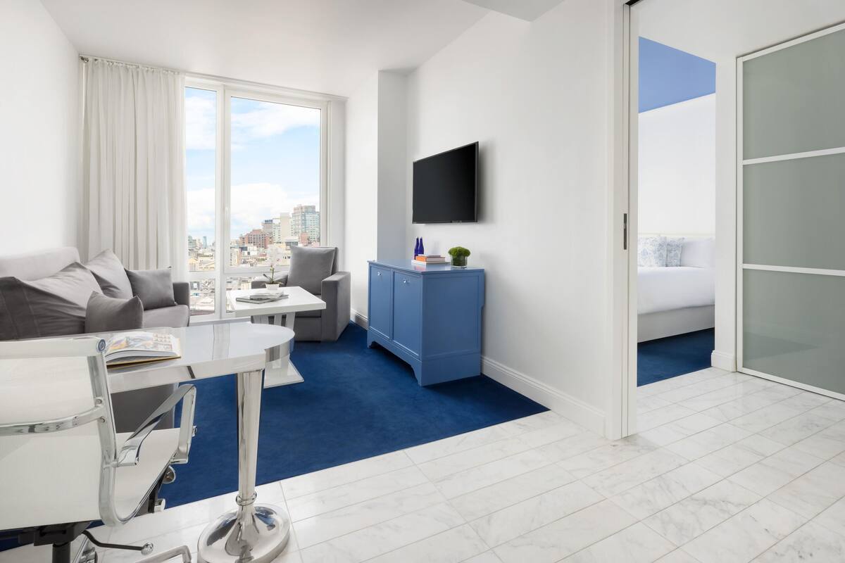 Une chambre d’hôtel moderne avec vue sur la ville, comprenant un lit, un bureau avec une chaise, une télévision et une moquette bleue.