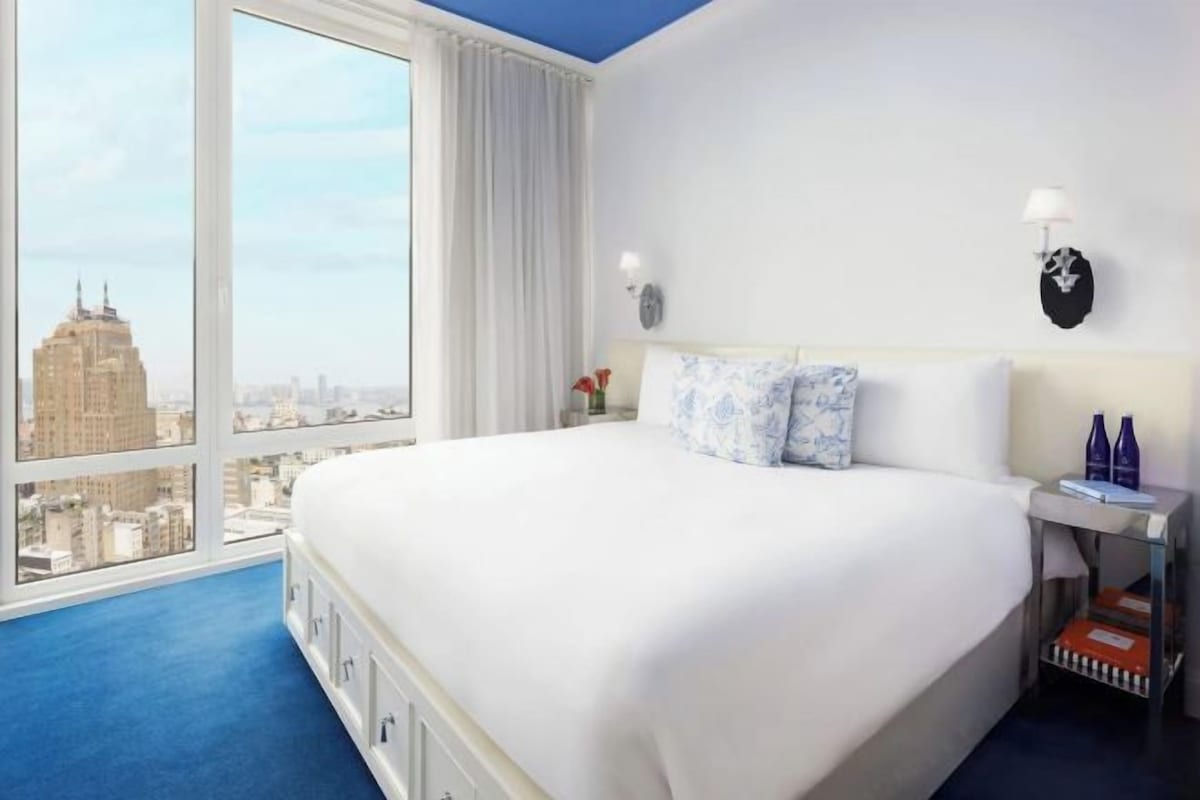 A hotel room with a large bed, a nightstand, a lamp, and a view of the cityscape.