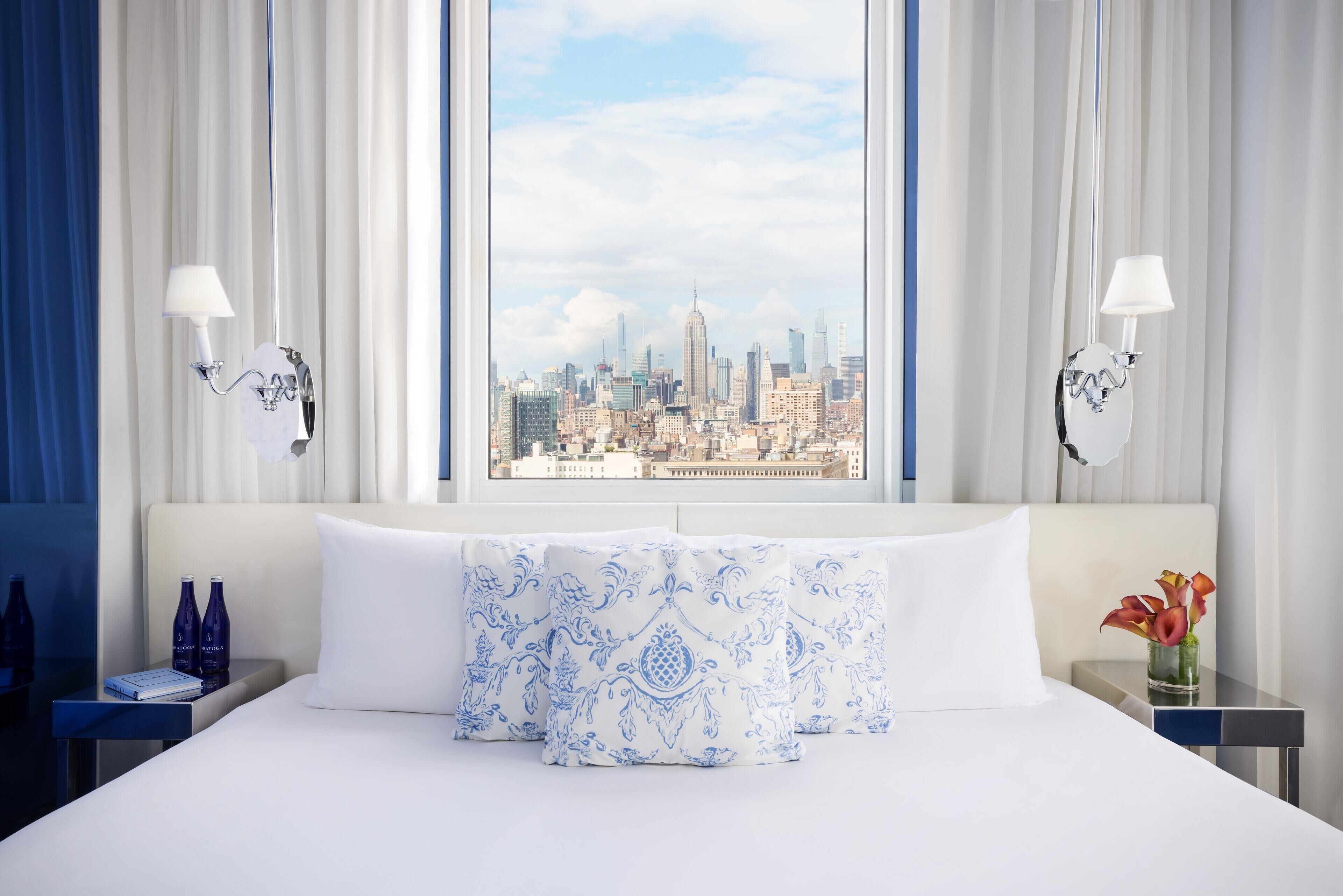 Habitación de hotel con una cama grande, dos mesitas de noche, una lámpara de noche, un ventanal con vistas a la ciudad y un jarrón con flores.