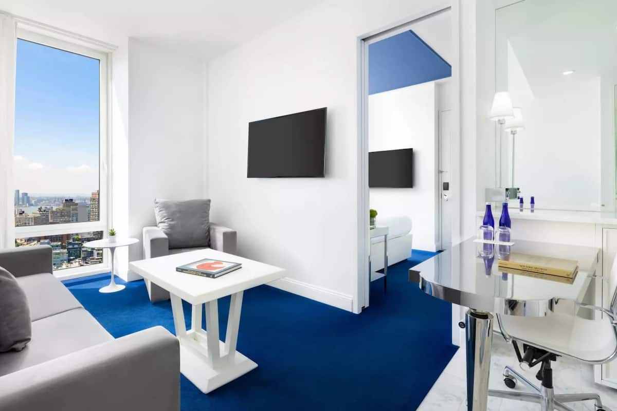 Une chambre d’hôtel moderne avec vue sur la ville, une télévision à écran plat, une petite table avec un livre, un canapé et une salle de bain équipée d’un miroir et d’articles de toilette.