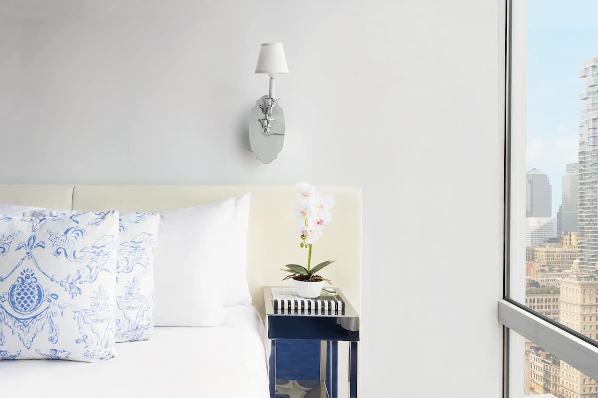 Une chambre d’hôtel avec un lit, une table de chevet ornée d’une orchidée en pot, une lampe fixée au mur et une vue sur la ville par la fenêtre.