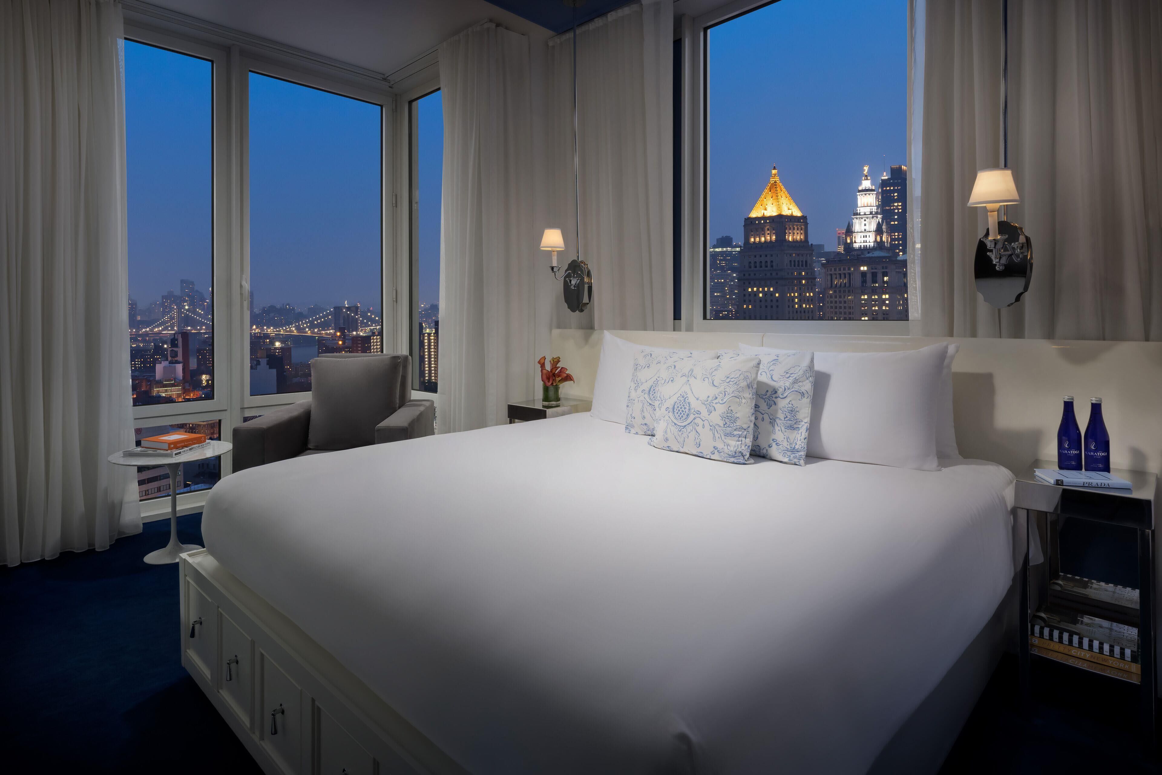 Una habitación de hotel con una cama grande, un escritorio con silla, una mesita de noche con lámpara, y vistas a una ciudad al anochecer.