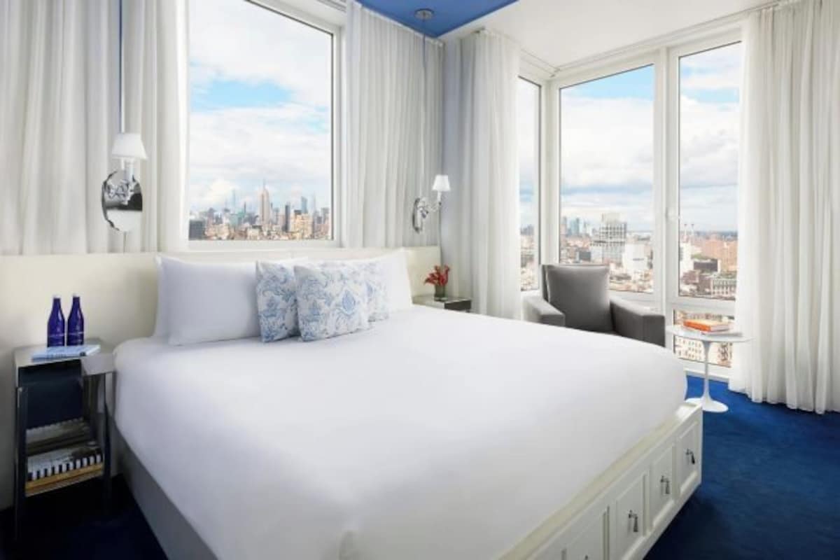 A hotel room with a large bed, two windows, a city view, a nightstand, and a chair.