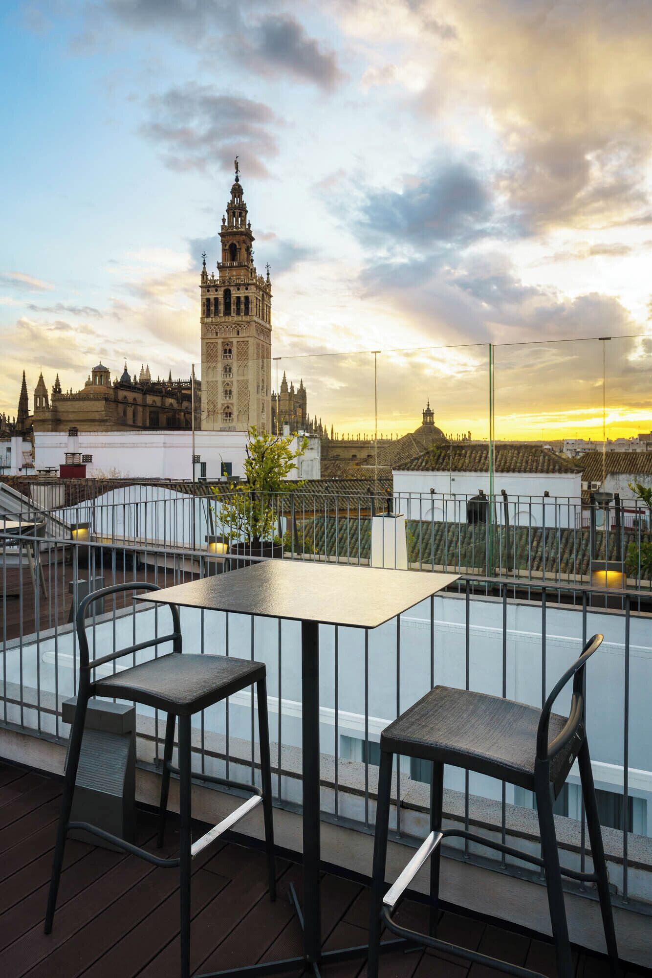 A rooftop terrace with two tables and chairs, overlooking a historic tower and cityscape during sunset.