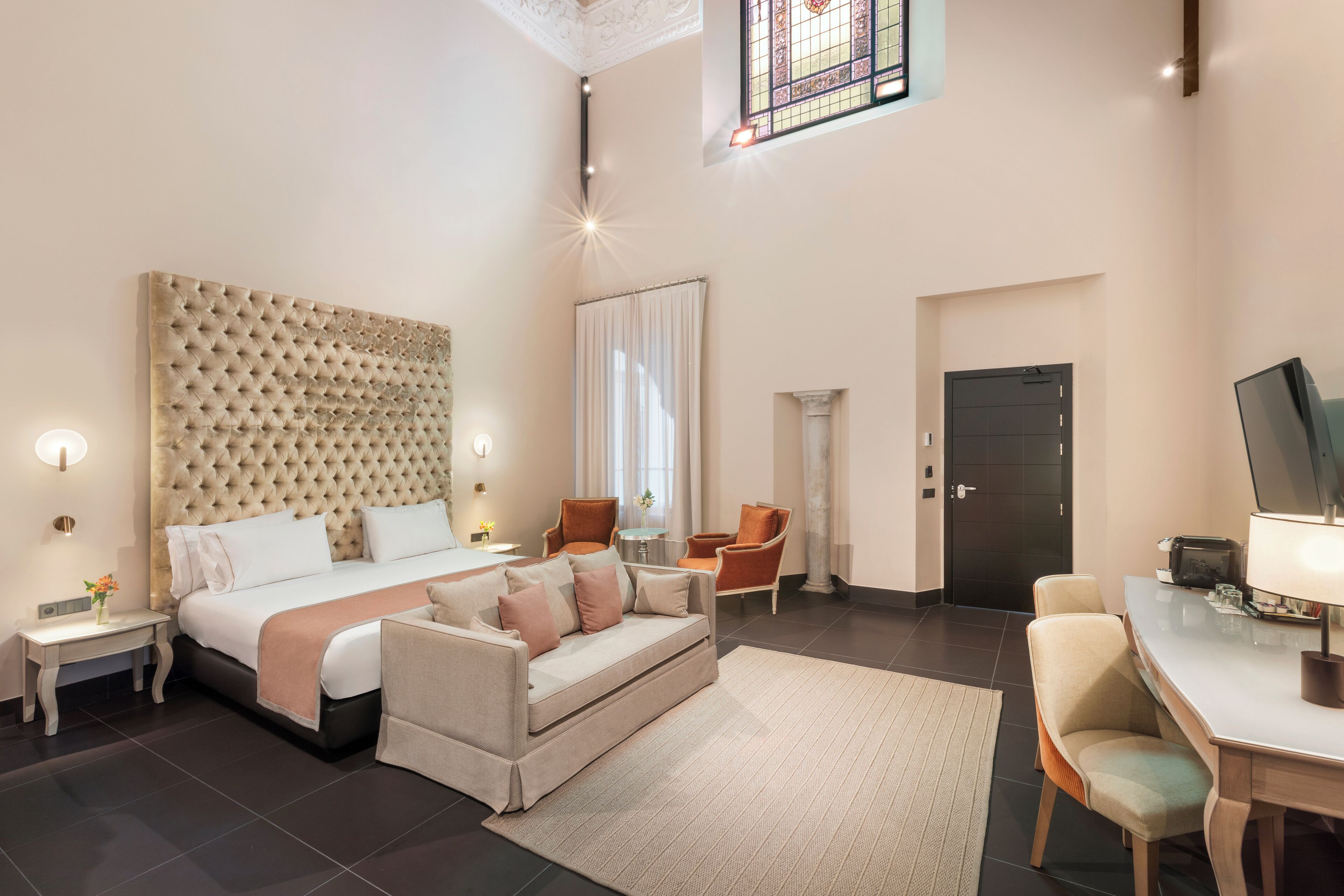 Une chambre d’hôtel moderne dotée d’une grande tête de lit capitonnée, d’un canapé, d’un bureau et d’une chaise.