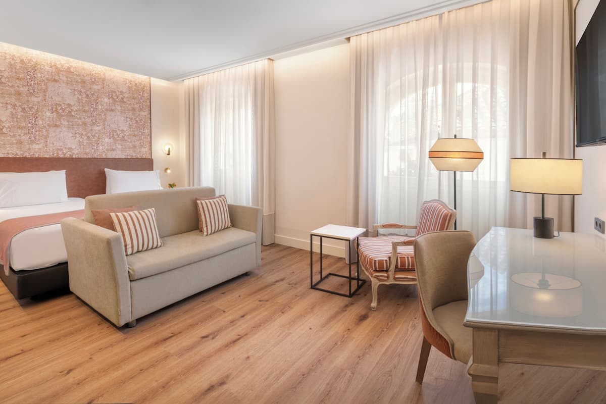 A hotel room with a bed, a sofa, a small table, a chair, and a desk with a lamp.