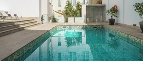 A swimming pool with a geometric tile pattern, a sun terrace with lounge chairs, and a building with windows and plants.