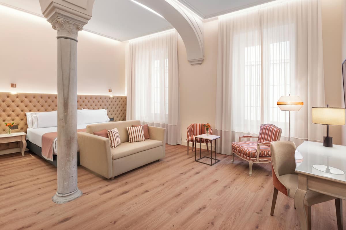 A hotel room with a tufted headboard bed, a beige sofa, a striped armchair, a small table with a lamp, and a large pillar.