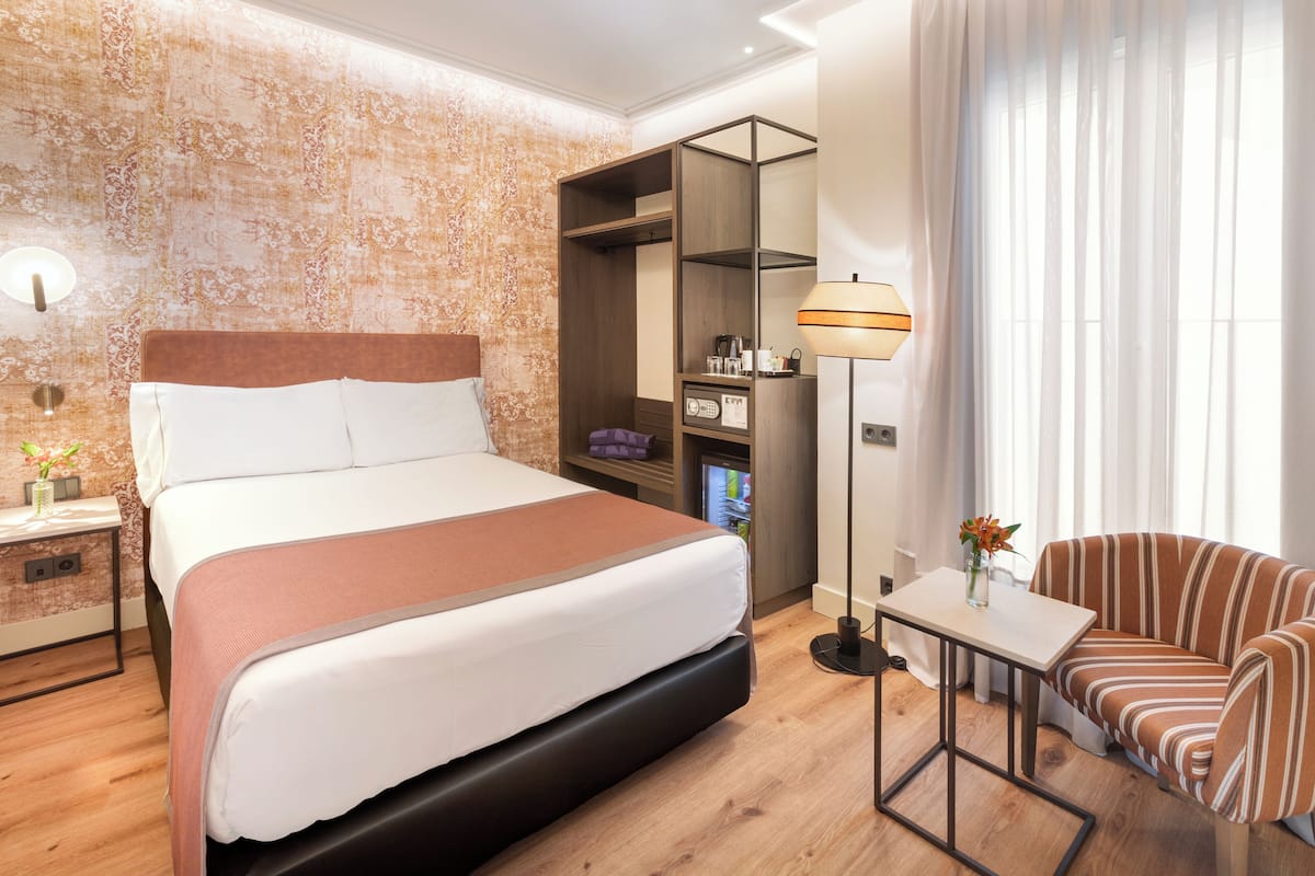 A modern hotel room with a large bed, a bedside table, a chair, a desk, a wardrobe, and a lamp.