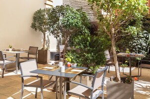An outdoor cafe with tables and chairs, surrounded by potted plants and trees.