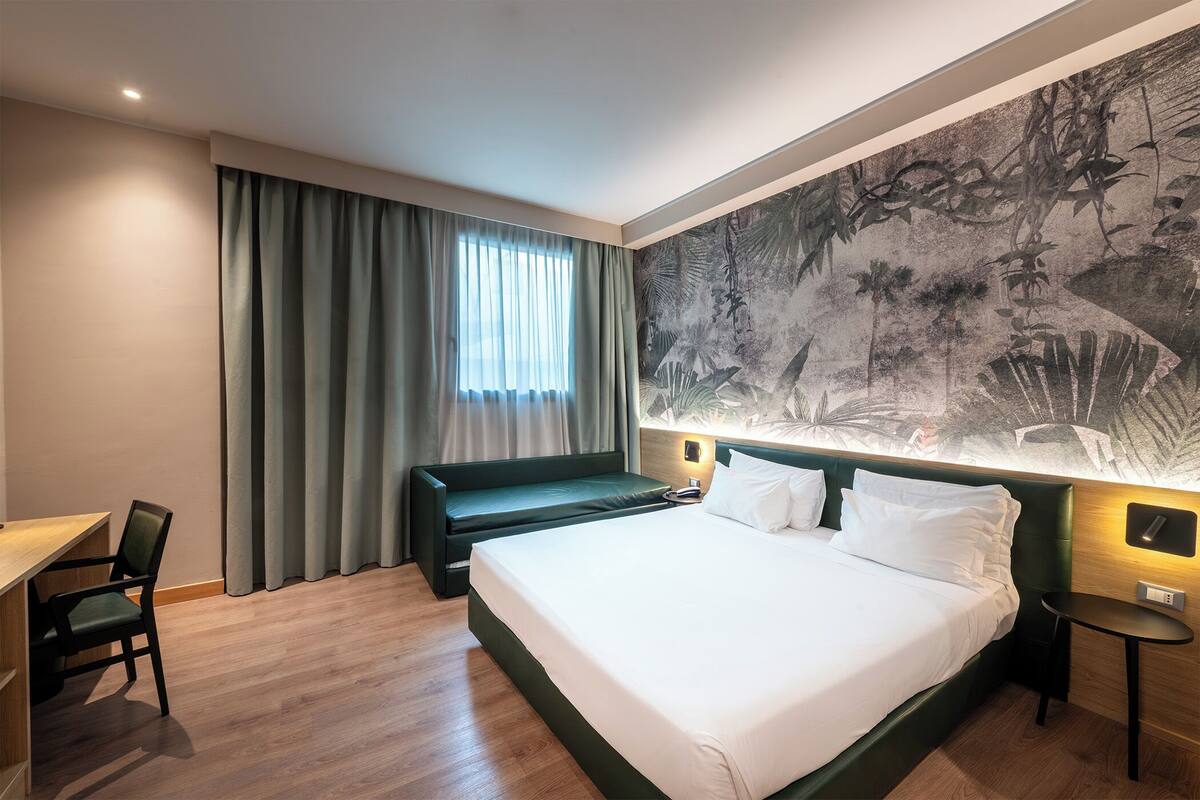 Hotellihuoneessa on suuri sänky, työpöytä ja tuoli. Huoneessa on ikkuna, jossa on verhot, seinämaalaus ja yöpöytä, jossa on lamppu.