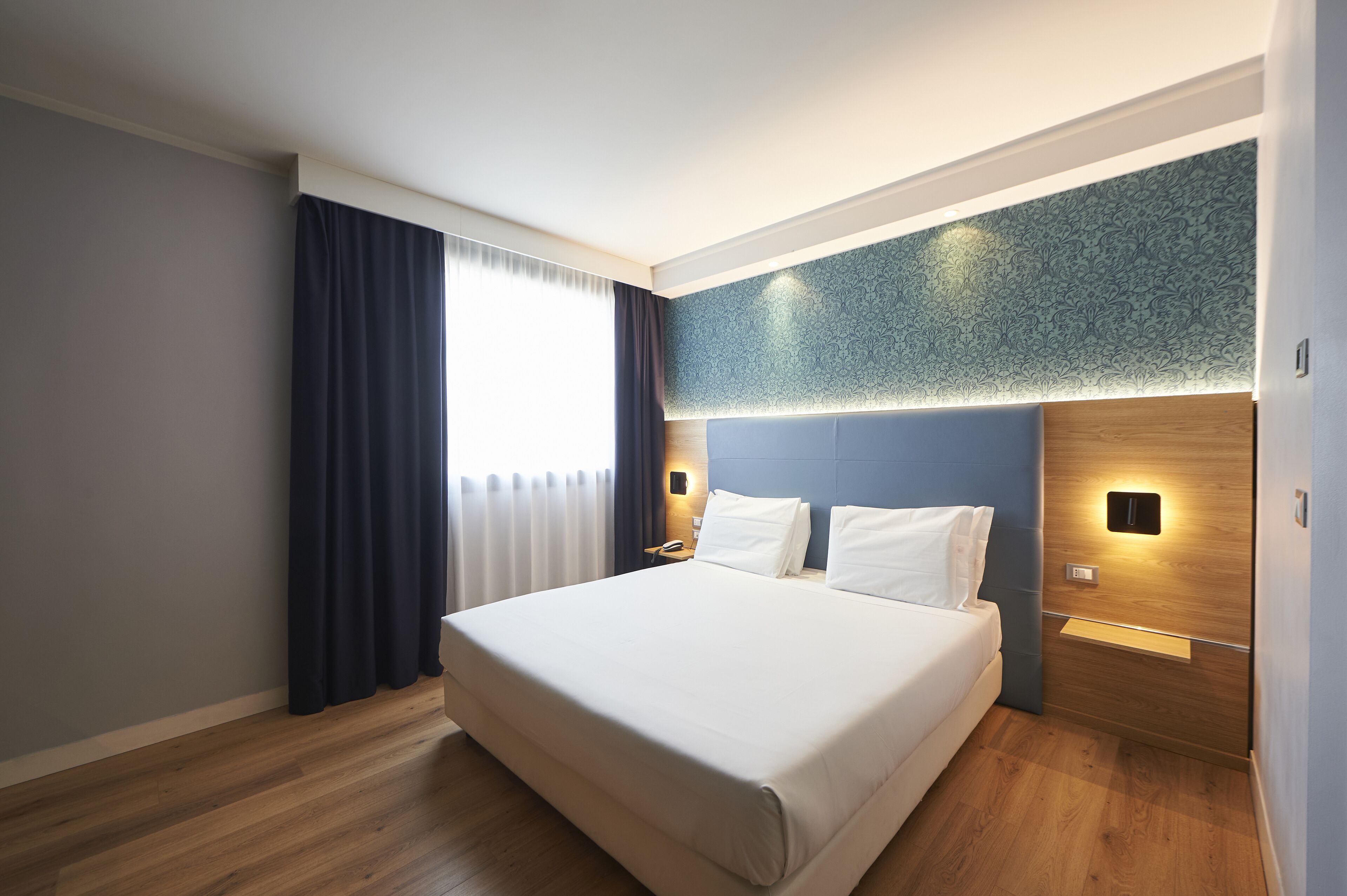 Habitación de hotel moderna con una cama grande, suelo de madera, una mesita de noche y una lámpara de pared.