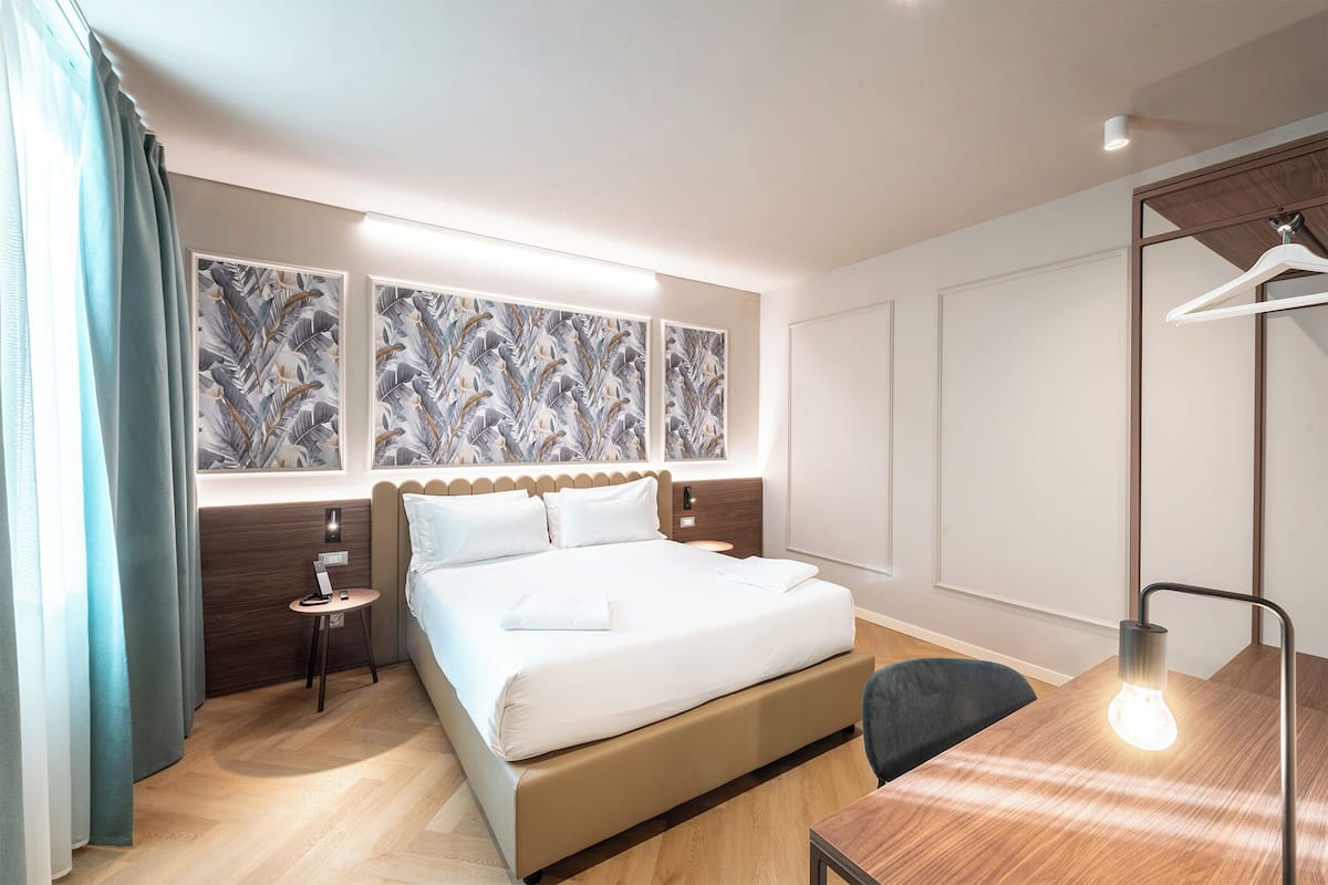 Moderni hotellihuone, jossa on suuri sänky, yöpöydät, työpöytä lampun kanssa ja taideteoksia seinillä.
