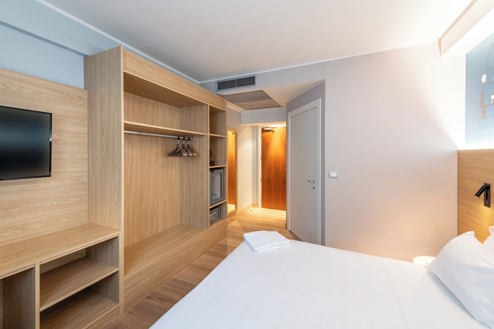 A modern hotel room with a wooden wardrobe, a bed, and a television.
