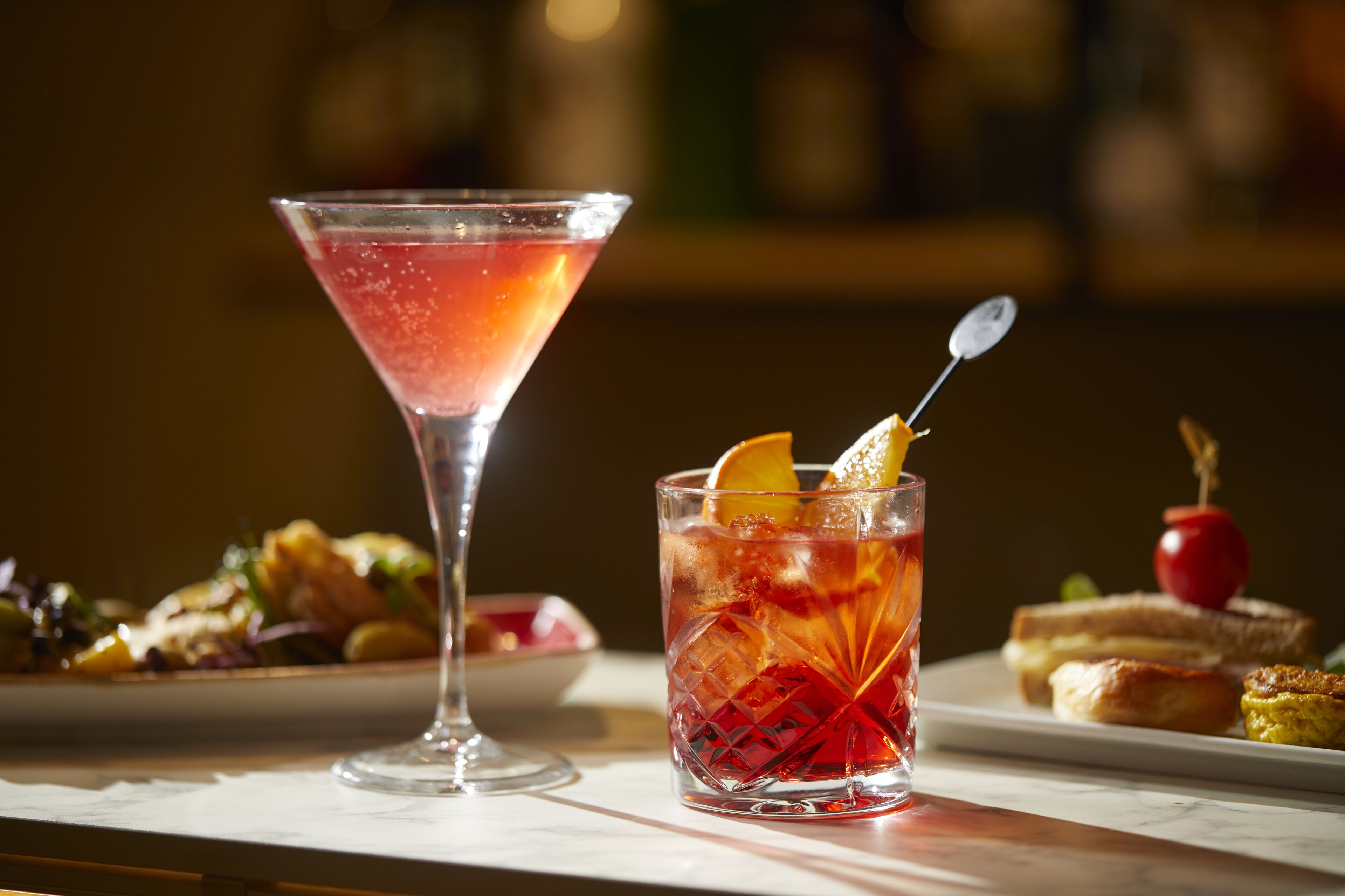 Un verre à martini et un verre à cocktail, tous deux décorés de fruits, sont posés sur une table où se trouvent également des plats.