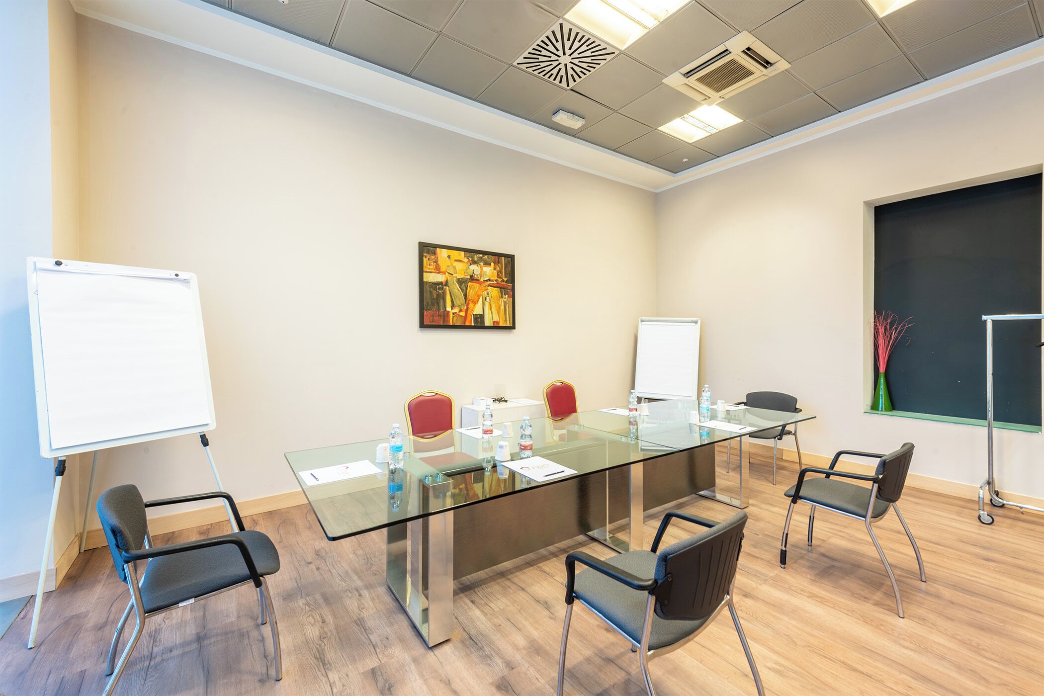 Un salón de conferencias con una mesa de vidrio, cuatro sillas, una pizarra blanca, una pizarra y un cuadro en la pared.