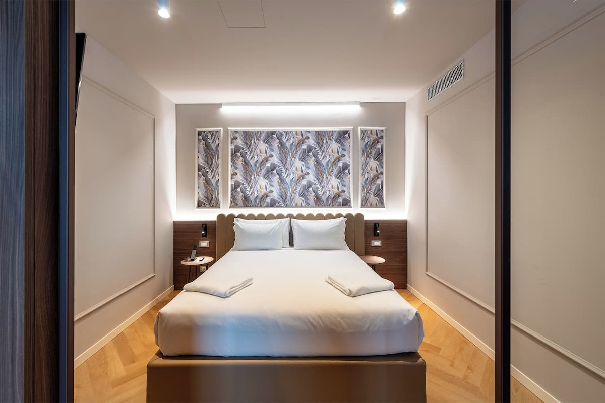Moderni hotellihuone, jossa on suuri sänky, kaksi yöpöytää, seinälle ripustettu taideteos ja puulattia.