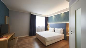 A modern hotel room with a wooden desk, a bed with white linens, a nightstand, and a large window with curtains.