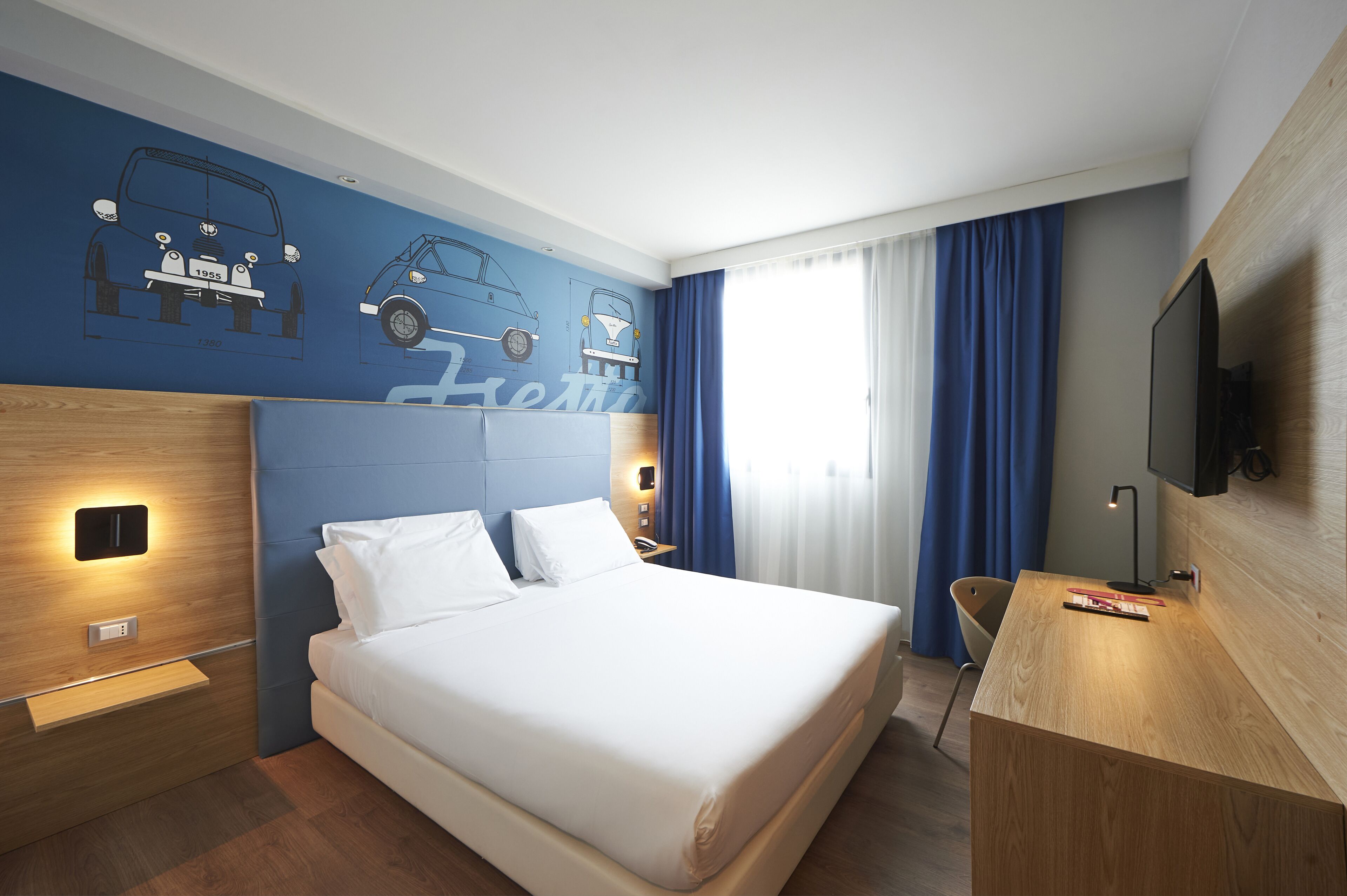 Une chambre d’hôtel avec un grand lit, un bureau, une télévision et un mur décoré d’illustrations de voitures.